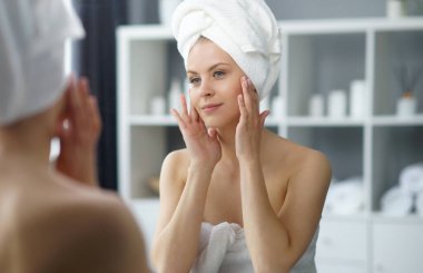 Young woman sits in the bathroom in front of the makeup mirror and does cosmetic procedures. Beautiful girl in white towel. The concept of skin care, health, rejuvenation and spa treatment.