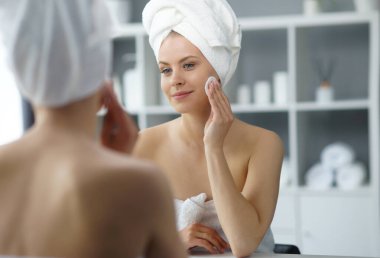 Young woman sits in the bathroom in front of the makeup mirror and does cosmetic procedures. Beautiful girl in white towel. The concept of skin care, health, rejuvenation and spa treatment.