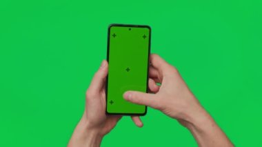 Male hand holding a smartphone with vertical green chroma key screen isolated on green background. Mobile phone in a hand. Different signs and gestures with fingers. 4K template. Technology and