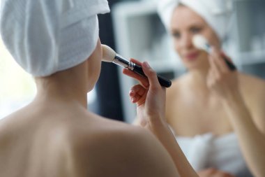 Young woman sits in the bathroom in front of the makeup mirror and does cosmetic procedures. Beautiful girl in white towel. The concept of skin care, health, rejuvenation and spa treatment.