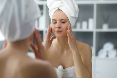 Young woman sits in the bathroom in front of the makeup mirror and does cosmetic procedures. Beautiful girl in white towel. The concept of skin care, health, rejuvenation and spa treatment.