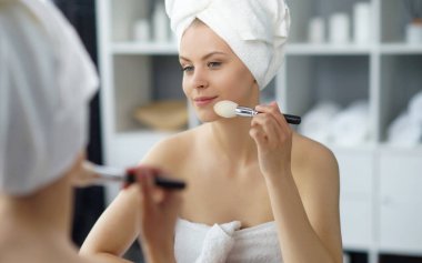 Young woman sits in the bathroom in front of the makeup mirror and does cosmetic procedures. Beautiful girl in white towel. The concept of skin care, health, rejuvenation and spa treatment.
