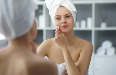 Young woman sits in the bathroom in front of the makeup mirror and does cosmetic procedures. Beautiful girl in white towel. The concept of skin care, health, rejuvenation and spa treatment.