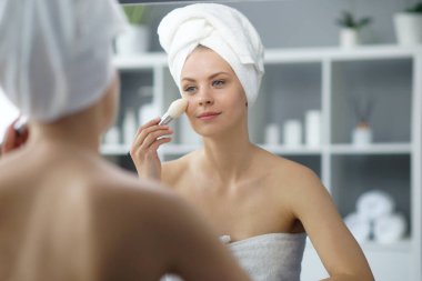 Young woman sits in the bathroom in front of the makeup mirror and does cosmetic procedures. Beautiful girl in white towel. The concept of skin care, health, rejuvenation and spa treatment.