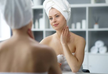 Young woman sits in the bathroom in front of the makeup mirror and does cosmetic procedures. Beautiful girl in white towel. The concept of skin care, health, rejuvenation and spa treatment.