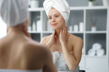 Young woman sits in the bathroom in front of the makeup mirror and does cosmetic procedures. Beautiful girl in white towel. The concept of skin care, health, rejuvenation and spa treatment.