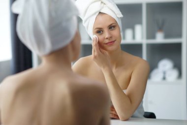 Young woman sits in the bathroom in front of the makeup mirror and does cosmetic procedures. Beautiful girl in white towel. The concept of skin care, health, rejuvenation and spa treatment.