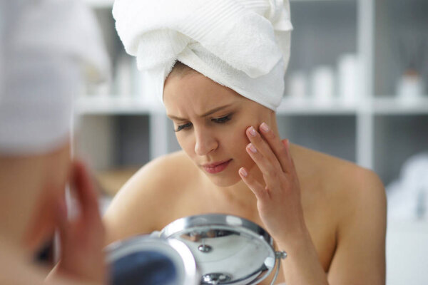 Young woman sits in the bathroom in front of the makeup mirror and does cosmetic procedures. Beautiful girl in white towel. The concept of skin care, health, rejuvenation and spa treatment.