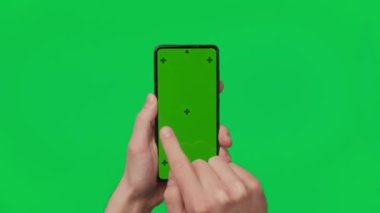 Male hand holding a smartphone with vertical green chroma key screen isolated on green background. Mobile phone in a hand. Different signs and gestures with fingers. 4K template. Technology and