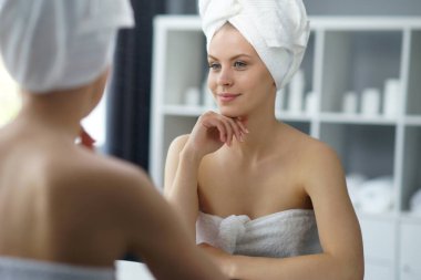 Young woman sits in the bathroom in front of the makeup mirror and does cosmetic procedures. Beautiful girl in white towel. The concept of skin care, health, rejuvenation and spa treatment.