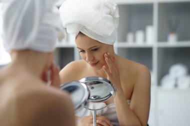 Young woman sits in the bathroom in front of the makeup mirror and does cosmetic procedures. Beautiful girl in white towel. The concept of skin care, health, rejuvenation and spa treatment.