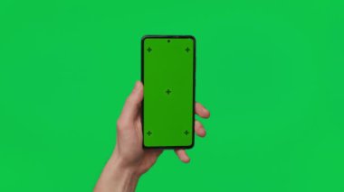 Male hand holding a smartphone with vertical green chroma key screen isolated on green background. Mobile phone in a hand. Different signs and gestures with fingers. 4K template. Technology and