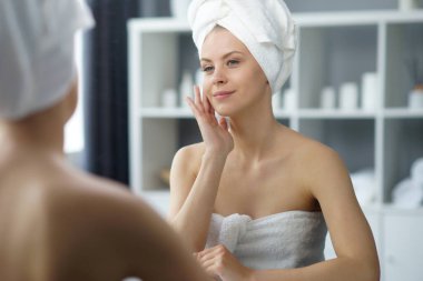 Young woman sits in the bathroom in front of the makeup mirror and does cosmetic procedures. Beautiful girl in white towel. The concept of skin care, health, rejuvenation and spa treatment.