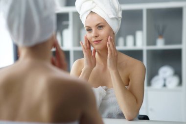 Young woman sits in the bathroom in front of the makeup mirror and does cosmetic procedures. Beautiful girl in white towel. The concept of skin care, health, rejuvenation and spa treatment.