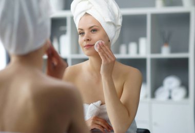 Young woman sits in the bathroom in front of the makeup mirror and does cosmetic procedures. Beautiful girl in white towel. The concept of skin care, health, rejuvenation and spa treatment.