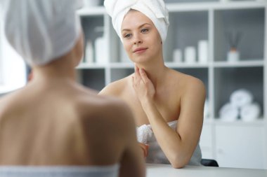 Young woman sits in the bathroom in front of the makeup mirror and does cosmetic procedures. Beautiful girl in white towel. The concept of skin care, health, rejuvenation and spa treatment.
