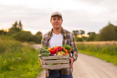 Gün batımında tarım arazisinin önünde sebze kutusu olan bir çiftçi. Kırsal kesimde bir adam. Kır hayatı, gıda üretimi, tarım ve kırsal yaşam tarzı kavramı.