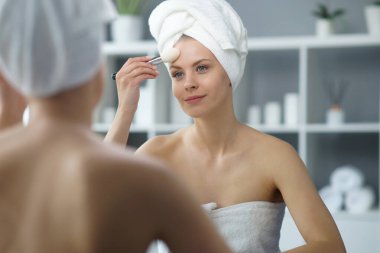 Young woman sits in the bathroom in front of the makeup mirror and does cosmetic procedures. Beautiful girl in white towel. The concept of skin care, health, rejuvenation and spa treatment.