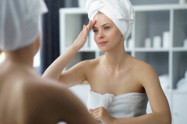Young woman sits in the bathroom in front of the makeup mirror and does cosmetic procedures. Beautiful girl in white towel. The concept of skin care, health, rejuvenation and spa treatment.