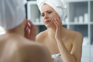 Young woman sits in the bathroom in front of the makeup mirror and does cosmetic procedures. Beautiful girl in white towel. The concept of skin care, health, rejuvenation and spa treatment.