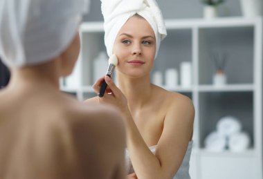 Young woman sits in the bathroom in front of the makeup mirror and does cosmetic procedures. Beautiful girl in white towel. The concept of skin care, health, rejuvenation and spa treatment.