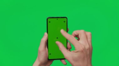 Male hand holding a smartphone with vertical green chroma key screen isolated on green background. Mobile phone in a hand. Different signs and gestures with fingers. 4K template. Technology and