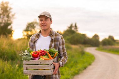 Farmer with a vegetable box in front of a sunset agricultural landscape. Man in a countryside field. The concept of country life, food production, farming and country lifestyle.