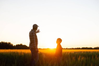 Farmer and his son in front of a sunset agricultural landscape. Man and a boy in a countryside field. The concept of fatherhood, country life, farming and country lifestyle.