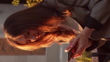 Genç çekici iş kadını dışarıda bankta oturuyor ve akıllı telefon kullanıyor. Gün batımı ışığı. Dikey video.