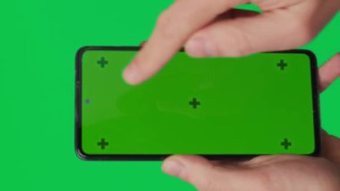 Yeşil arka planda izole edilmiş dikey yeşil renkli anahtar ekranlı akıllı telefonu tutan bir erkek. Cep telefonu bir elde. Farklı işaretler ve parmaklarla yapılan jestler. 4K dikey video şablonu