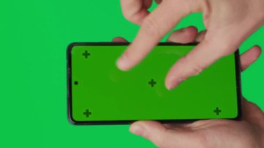 Yeşil arka planda izole edilmiş dikey yeşil renkli anahtar ekranlı akıllı telefonu tutan bir erkek. Cep telefonu bir elde. Farklı işaretler ve parmaklarla yapılan jestler. 4K dikey video şablonu