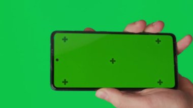 Yeşil arka planda izole edilmiş dikey yeşil renkli anahtar ekranlı akıllı telefonu tutan bir erkek. Cep telefonu bir elde. Farklı işaretler ve parmaklarla yapılan jestler. 4K dikey video şablonu