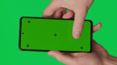 Yeşil arka planda izole edilmiş dikey yeşil renkli anahtar ekranlı akıllı telefonu tutan bir erkek. Cep telefonu bir elde. Farklı işaretler ve parmaklarla yapılan jestler. 4K video şablonu