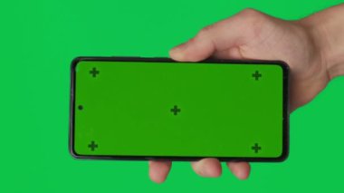 Yeşil arka planda izole edilmiş dikey yeşil renkli anahtar ekranlı akıllı telefonu tutan bir erkek. Cep telefonu bir elde. Farklı işaretler ve parmaklarla yapılan jestler. 4K video şablonu