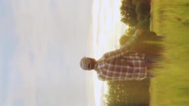 Çiftçi ve oğlu günbatımı tarımının önünde. Kırsalda bir adam ve bir çocuk. Babalık kavramı, köy hayatı, çiftçilik ve kırsal yaşam tarzı. Dikey video.