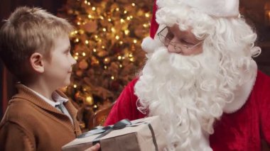 Noel Baba bir masada oturuyor, küçük bir çocukla mutlu bir şekilde çalışıyor. Arka planda, sıcak bir şömine ve güzel süslenmiş bir Noel ağacı klasik bir tatil atmosferi yaratır..