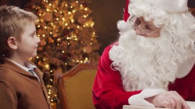 Noel Baba bir masada oturuyor, küçük bir çocukla mutlu bir şekilde çalışıyor. Arka planda, sıcak bir şömine ve güzel süslenmiş bir Noel ağacı klasik bir tatil atmosferi yaratır..