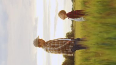 Çiftçi ve oğlu günbatımı tarımının önünde. Kırsalda bir adam ve bir çocuk. Babalık kavramı, köy hayatı, çiftçilik ve kırsal yaşam tarzı. Dikey video.