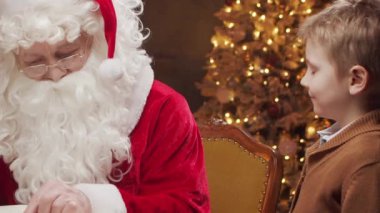 Noel Baba bir masada oturuyor, küçük bir çocukla mutlu bir şekilde çalışıyor. Arka planda, sıcak bir şömine ve güzel süslenmiş bir Noel ağacı klasik bir tatil atmosferi yaratır..