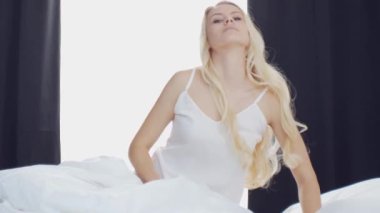 Yatakta uzanan genç kadın, büyük perdelerin arasından süzülen güneş ışığıyla çerçevelenmiş, sabah dinginliği ve gençleşme hissi uyandırıyor. Ev kavramı.