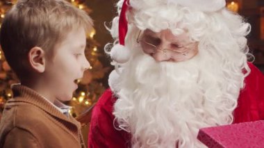 Noel Baba ve küçük çocuk. Neşeli Noel Baba masada otururken çalışıyor. Arka planda şömine ve Noel ağacı var. Geleneksel Noel kavramı.