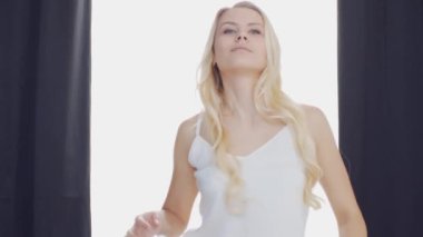 Yatakta uzanan genç kadın, büyük perdelerin arasından süzülen güneş ışığıyla çerçevelenmiş, sabah dinginliği ve gençleşme hissi uyandırıyor. Ev kavramı.