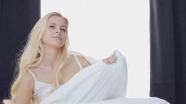 Yatakta uzanan genç kadın, büyük perdelerin arasından süzülen güneş ışığıyla çerçevelenmiş, sabah dinginliği ve gençleşme hissi uyandırıyor. Ev kavramı.