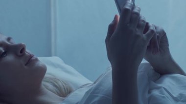 Genç bir kadın yatakta uzanıyor, rahat bir şekilde akıllı telefon kullanıyor, rahat bir sabah rutini uyguluyor. Yumuşak ışıklandırma ve rahat ortam bağlantı ve modern yaşam tarzının temalarını vurguluyor.