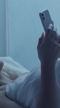 Genç bir kadın yatakta uzanıyor, rahat bir şekilde akıllı telefon kullanıyor, rahat bir sabah rutini uyguluyor. Yumuşak ışıklandırma ve rahat ortam, bağlantı ve modern yaşam tarzının temalarını vurguluyor. Dikey video.
