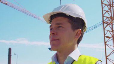 İnşaat alanının önünde duran profesyonel müteahhit. Kasklı ve yelekli ustabaşı. Ofis binası ve vinç arka planı. İş, gayrimenkul ve yatırım kavramları.