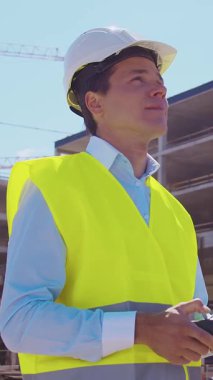 İnşaat alanının önünde duran profesyonel müteahhit. Kasklı ve yelekli ustabaşı. Ofis binası ve vinç arka planı. İş, gayrimenkul ve yatırım kavramları.