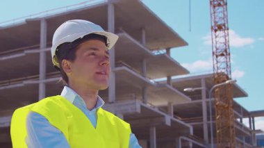 İnşaat alanının önünde duran profesyonel müteahhit. Kasklı ve yelekli ustabaşı. Ofis binası ve vinç arka planı. İş, gayrimenkul ve yatırım kavramları.