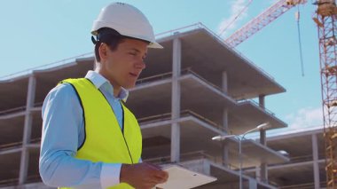 İnşaat alanının önünde duran profesyonel müteahhit. Kasklı ve yelekli ustabaşı. Ofis binası ve vinç arka planı. İş, gayrimenkul ve yatırım kavramları.