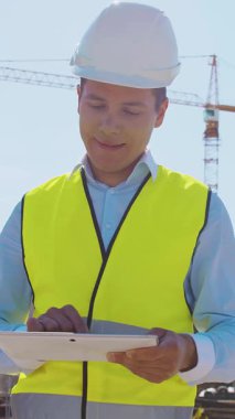 İnşaat alanının önünde duran profesyonel müteahhit. Kasklı ve yelekli ustabaşı. Ofis binası ve vinç arka planı. İş, gayrimenkul ve yatırım kavramları.